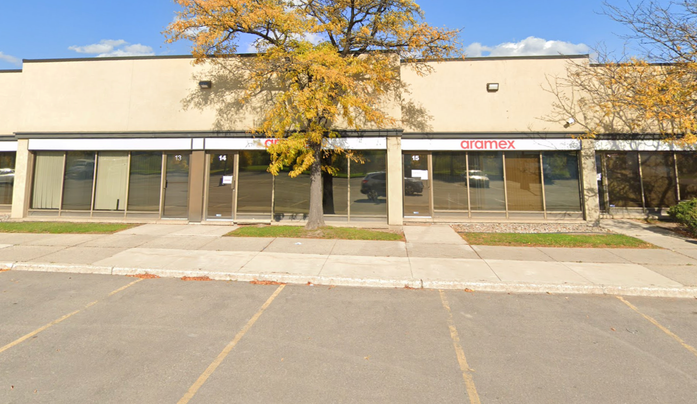 A one-story commercial building with large windows, a tree in front, and a sign reading "aramex." There are two glass doors labeled 14 and 15, and an empty parking lot in the foreground.