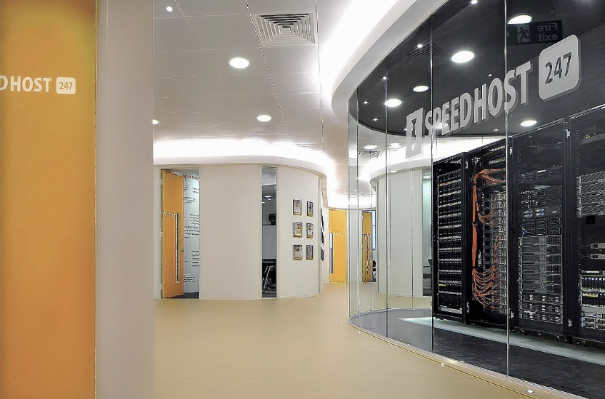 A modern server room with glass walls displays rows of network servers and cables. The space is brightly lit with clean white ceilings and yellow walls. Signs read "SPEEDHOST 247.