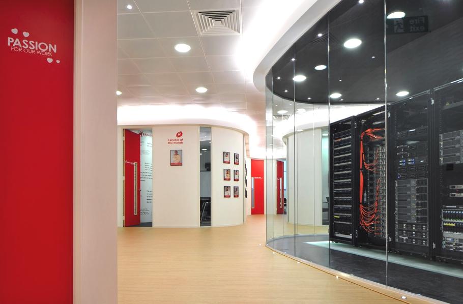 A modern office interior with red and white walls, motivational text saying "PASSION FOR OUR WORK," framed pictures, and a glass-walled server room with visible network equipment and cables.