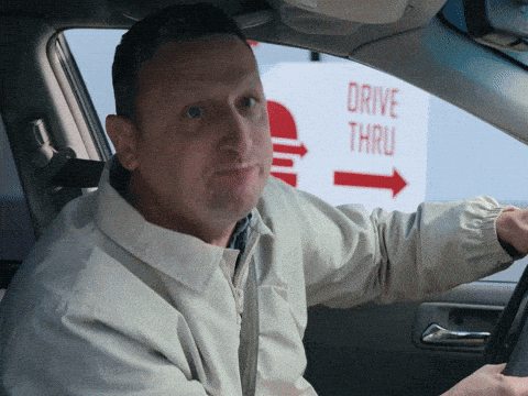 A man sitting in a car at a drive-thru looks surprised and mouths something, gesturing with his hand. A "Drive Thru" sign with a burger and arrow is visible outside the car window.