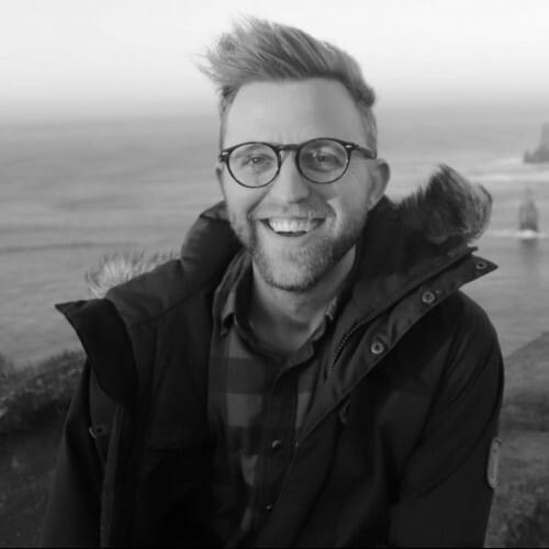 A smiling man with glasses wearing a jacket, sitting outdoors with a blurred coastal background during dusk.