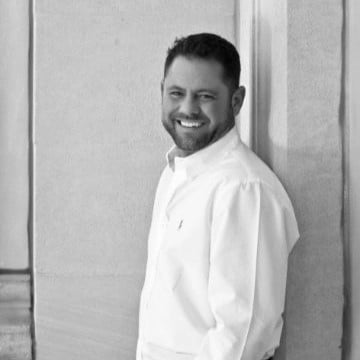 A smiling man in a white shirt leaning against a wall in a black and white photo.