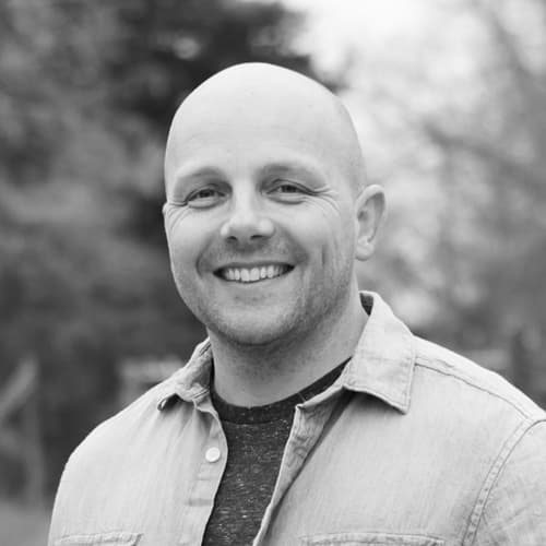 Black and white portrait of a smiling bald man wearing a shirt over a t-shirt, standing outdoors with trees blurred in the background.