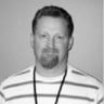 Black and white photo of a smiling man with a beard, wearing a striped shirt and a badge on a lanyard.