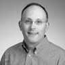 A black-and-white headshot of a middle-aged man smiling gently at the camera. he is balding, wears glasses, and is dressed in a checkered shirt.