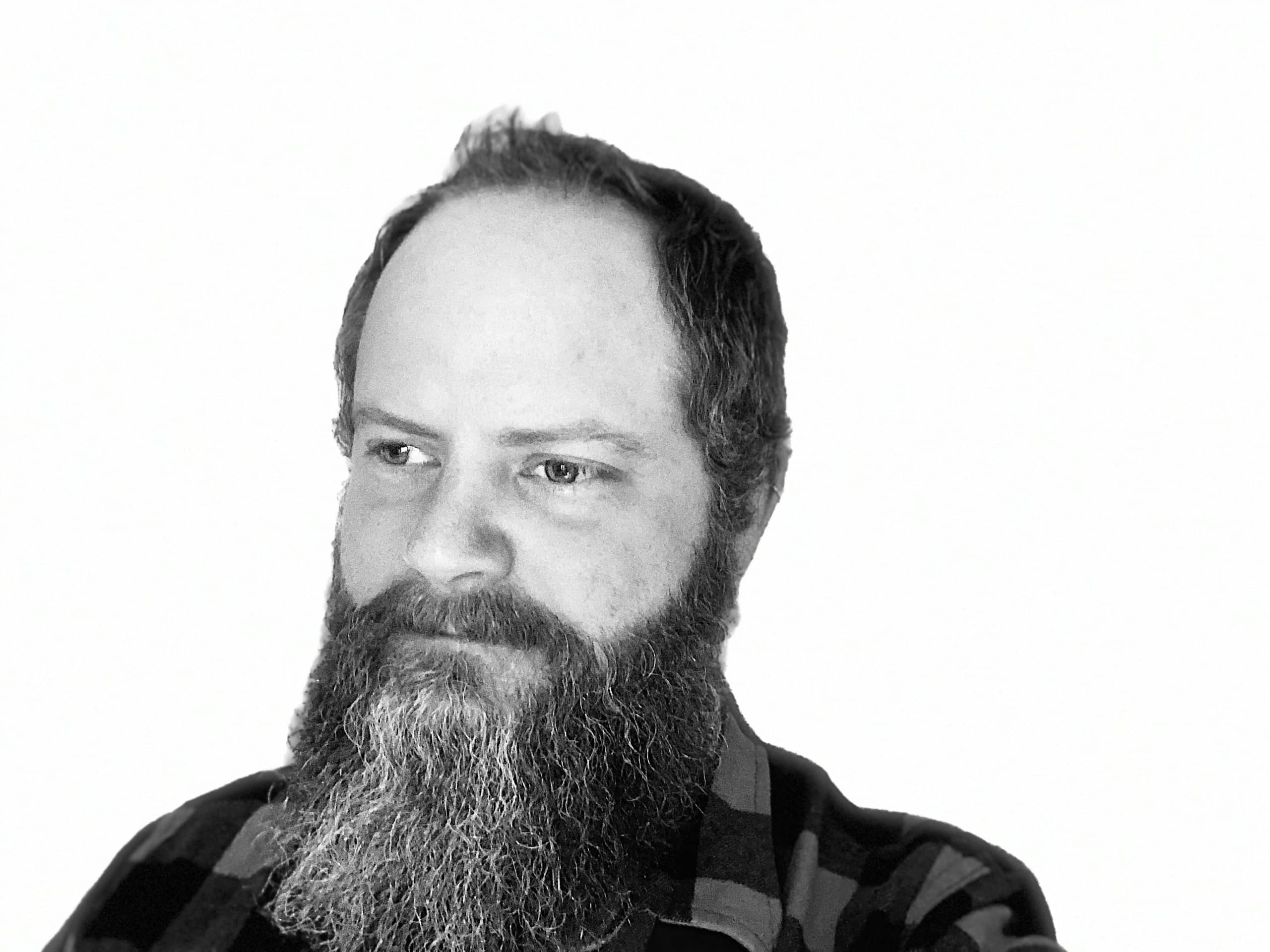 Black-and-white photo of a bearded man with short hair, wearing a plaid shirt, looking slightly to the side against a plain white background.