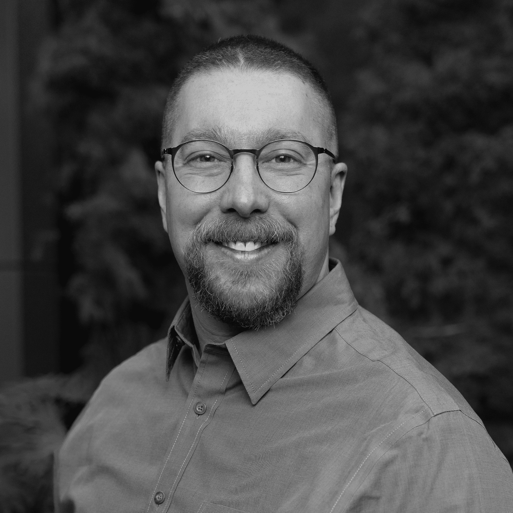 Man with short hair and glasses smiling, wearing a button-up shirt. The background is blurred with dark foliage. Image is in black and white.