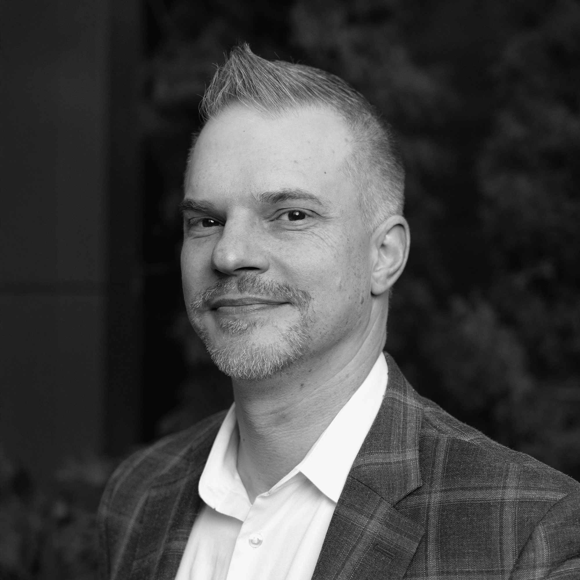 Black and white image of a person with short hair and a mustache, wearing a plaid blazer and a white shirt. They are looking slightly to the side with a neutral expression. The background is blurred with foliage.