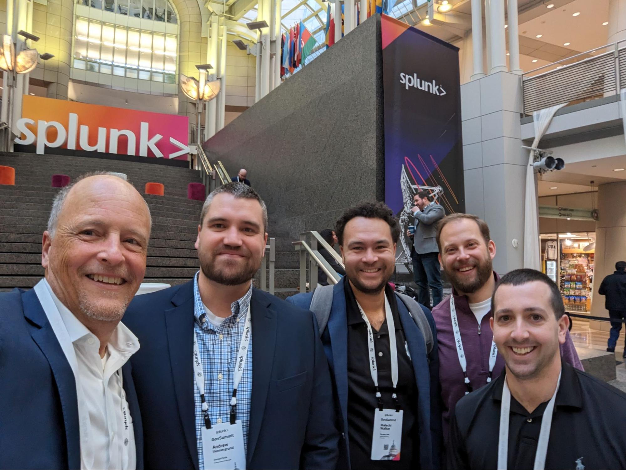Five smiling men wearing conference badges pose for a group photo inside a large convention center. Colorful "Splunk" signs and flags set the scene as they discuss navigating the world of AI and Zero Trust by tall staircases in the background.