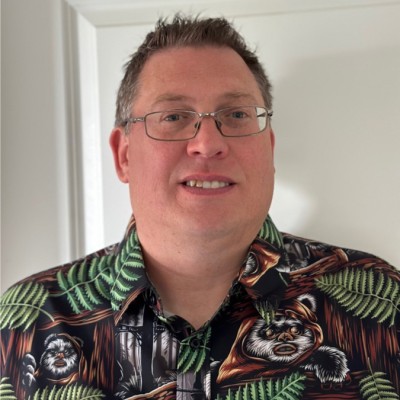 A man with short brown hair and glasses smiles at the camera. He is wearing a shirt with a pattern of ferns and cartoon-like animal faces on it, standing indoors in front of a white door.