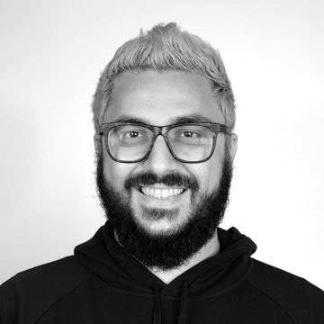 A black and white portrait of a smiling man with glasses and a full beard, wearing a hoodie. the background is plain and light-colored.