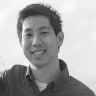 A black and white portrait of a smiling asian man with short hair, wearing a polo shirt, standing against a plain background.